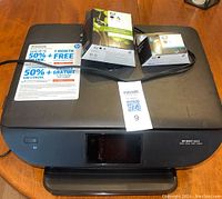 HP Envy 5660 printer on a wooden surface with visible scratches on the top cover, includes ink cartridge packaging and installed ink cartridges.