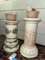 Two ceramic decorative columns side by side, each holding a ceramic bowl with round decorative balls inside. Columns have detailed floral and leaf designs in cream, green and red.