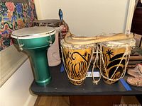 Photo showing three drums, two wooden with painted designs and natural skin heads, one green metal or fiberglass drum with white head, placed on a black surface with patterned textile background.