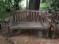 Front view of a weathered wooden garden bench set outdoors on a natural ground with pine needles and some greenery around. Bench has a curved top backrest with vertical slats and wooden armrests.