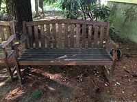 Front view of wooden garden bench placed outdoors on dirt ground with some foliage in background, showing vertical slats and armrests.