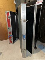 Two metal framed guitar cases standing upright. One case has silver metal exterior and black foam interior visible. The other is red with metal edges.