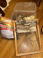 Full view of wooden lap loom with partially woven wool yarn textile and large plastic bin of wool skeins behind it plus a Hefty storage bag with crafting material.