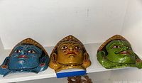 Front view of three colorful decorative masks on a white shelf, showing blue, yellow and green masks with detailed facial paint and gold colored headdresses.