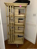 Full view of the wooden revolving bookshelf on wheels, showing vertical slat sides, four shelves filled with books (not included), and caster wheels at the bottom.