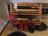 Wooden spice rack labeled Spice Islands in light wood finish, with pepper mill, salt and pepper shakers, metal container, and colorful nut chopper in front on stovetop.