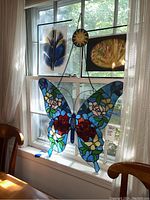 Photo showing stained glass butterfly suncatcher hanging in the window with sunlight illuminating colors, ceramic clay sun visible on chain above it, and two framed pictures in the background.