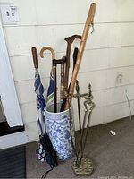 Wide view of ceramic umbrella stand filled with umbrellas, walking canes and brass fireplace tools in front of shiplap wall