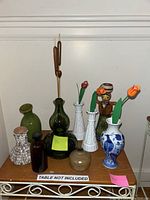 Full view of all vases on a table including green ceramic, dark green glass, white milk glass, blue and white porcelain, brown speckled, amber glass bottle, and owl-shaped vase.