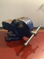 Front-side view of a small blue metal bench vise placed on a reddish surface, showing the serrated jaws and metal tightening handle.