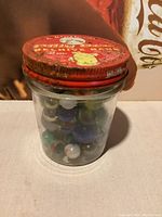 Front view of glass jar with red metal lid labeled 'Billy Bee Beehive Bank' filled with multicolored old marbles inside.