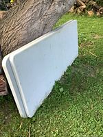 Two white plastic topped folding tables stacked leaning against a tree outdoors on grass.
