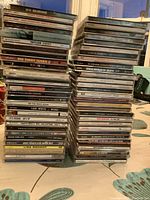 Three stacks of various music CD jewel cases showing multiple artists and album titles stacked on a surface.