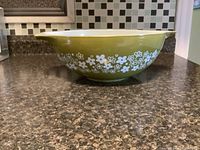 Side view of large olive green Pyrex mixing bowl on kitchen counter, showing white floral band around bowl exterior and pouring spout.