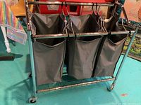 Front angle showing all three grey fabric bags suspended from a shiny chrome metal frame with wheels on a turquoise floor.