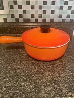 Side view of vintage orange Le Creuset sauce pot with lid on granite countertop, showing handle and lid knob.
