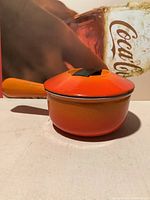 Side view of small flaming orange Le Creuset sauce pot with lid and handle.