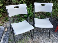 Two white solid Lifetime folding chairs with black metal frames placed on gravel outside, showing front and side views.