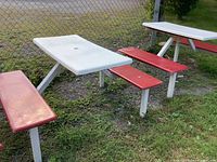 Two white rectangular outdoor picnic tables with attached red benches, placed on grass next to a chain-link fence, showing weathering and wear.