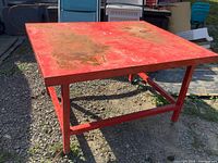Heavy, square red metal table with flat top and rust patches. Sturdy metal frame with horizontal support bars and leveling feet for stability.