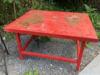 Complete view of top and legs of red metal table with rust spots and dirt, showing outdoor weathering.