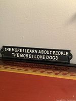 Front view of black metal sign with white raised text 'THE MORE I LEARN ABOUT PEOPLE THE MORE I LOVE DOGS'.