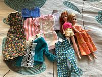 Two small plastic dolls and assorted old doll clothes laid out on a patterned surface, showing various garments and the dolls themselves.