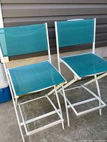 Two turquoise folding high patio chairs with white metal frames and mesh fabric seats and backs shown outside against house siding.