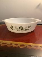 Side view of white oval milk glass casserole dish with green floral and leaf decorations along sides, placed on wooden surface.