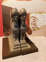 Front view of paired marble bookends showing human-headed winged lion figures mounted on stepped bases and brown rectangular platforms, standing side by side.