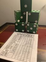 Photo of five large green wooden dice stacked in a pyramid with a Yardzee score card in front displaying different scoring categories and columns for players.