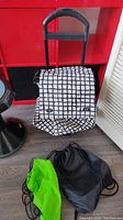 The set includes one black and white grid pattern rolling bag with handle and wheels, accompanied by two drawstring bags, one green and one black, placed on a floor near a red and black backdrop.