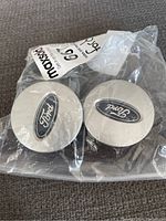 Two Ford wheel centre caps in sealed plastic bag, silver with blue oval logo.