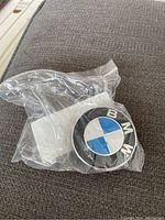 BMW center cap in original plastic bag placed on dark gray fabric, showing front logo facing up.