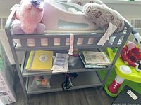 Wooden baby change table with gray frame, contoured white changing pad on top, two shelves with baby items such as books and small boxes. Positioned next to a window and other baby gear.