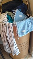 Cardboard packing barrel full of assorted clothing with visible denim shorts and striped shirt draped over the edge.