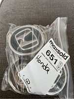 Four Honda center caps in a clear plastic zip bag with a MaxSold auction tag. Caps showcase the silver Honda H emblem with dark background and silver rim.