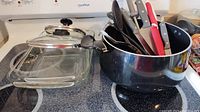 Two clear glass baking dishes, one covered with a glass lid. Large stainless steel pot holding multiple kitchen utensils including knives, a slotted spoon, a corkscrew, scissors, and a potato masher, all on a black stovetop.