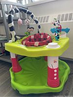 Full view of Evenflo infant saucer showing green base, pink and white legs, padded red and black checkered seat with hanging toys on overhead bar.