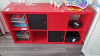 Red laminate shelving unit with six cubbies, two with black doors and one with red door. One red door is missing and one door pull is missing. The unit is shown from the front with a grey floor.