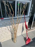 Full view of a tall clear glass vase on a patio floor against a brick wall with several tall decorative wood sticks inside, measuring about 60 inches tall.