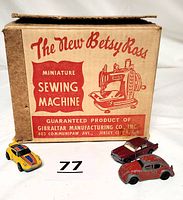 Original cardboard box of the Betsy Ross miniature sewing machine alongside three vintage diecast cars: red Volkswagen Beetle, yellow Hot Wheels Porsche Carrera, and silver Rolls Royce Silver Shadow with trunk lid missing.