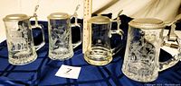 Front view of four etched clear glass beer steins with pewter lids arranged on dark cloth
