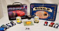 Full lot view showing McQueen Lightning 95 metal lunch box, Bingo game tin with cards and chip dispenser, two Simpsons figures, Garfield eraser packaging, and metal cars.