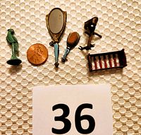 Overview of four vintage miniature dollhouse accent pieces including metal parakeet, hand mirror and brush, stereoscope viewer, and baby doll bed laid on textured white fabric next to a penny for scale.