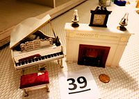 White miniature dollhouse grand piano with lid open showing artwork on underside, matching red cushioned bench, miniature sheet music, and black metronome next to a white miniature fireplace with a small clock and two lamps on mantel. A penny placed for scale.