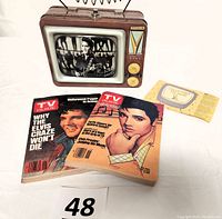 Front view of the Elvis Presley Vandor tin lunch box shaped like a TV, showing the black and white Elvis image, antenna handle, dials, and the two TV Guide magazines in front dated 1963 and 1995.
