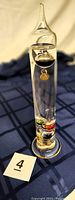 Front view of the 13-inch Galileo thermometer showing the clear glass body with colored glass balls suspended inside and a small number tag for each ball. The item is standing on a blue patterned surface.