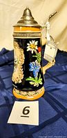 Side view of the beer stein showing detailed floral decorations in bright blue, white, and red colors with attached tag on handle.