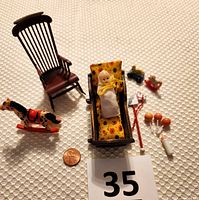 Lot of miniature vintage dollhouse toys arranged on white textured fabric including rocking horse, cradle with baby doll, rocking chair, stick hobby horse, duck pull toy, baby bottle, and rattles along with a penny for scale.